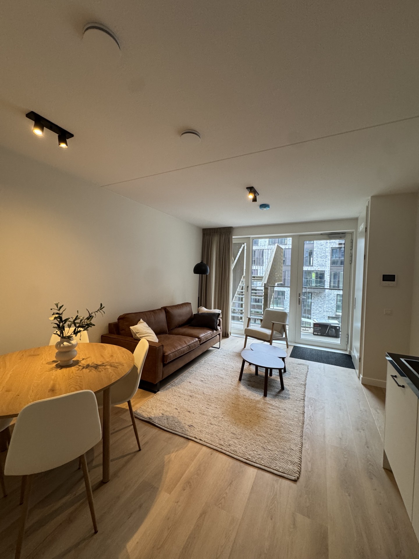 Lichte woonkamer met cognackleurige leren bank, ronde eettafel in eiken, zwarte vloerlamp en doorkijk naar het balkon, voorbeeld van een compleet ingerichte ruimte door RAW-Building