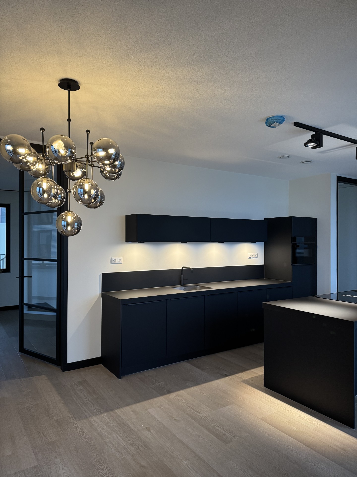 Zwarte keuken met glazen bollamp, zwart kookeiland en houten visgraatvloer